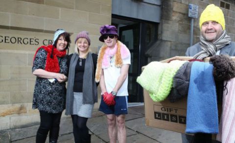 A man carrying a box of warm clothes and blankets in the foreground, with three women wearing scarves and blankets in the background.