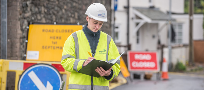 A Northern Gas Networks employee wearing a hi-vis vest and safety headwear, writing in a notebook. Various roadside construction equipment is in the background.