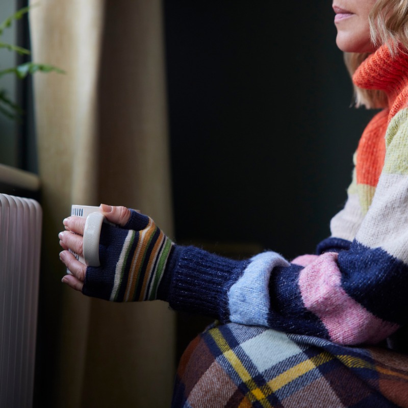 Image of person sat in front of radiator with a cup of tea in their hand.