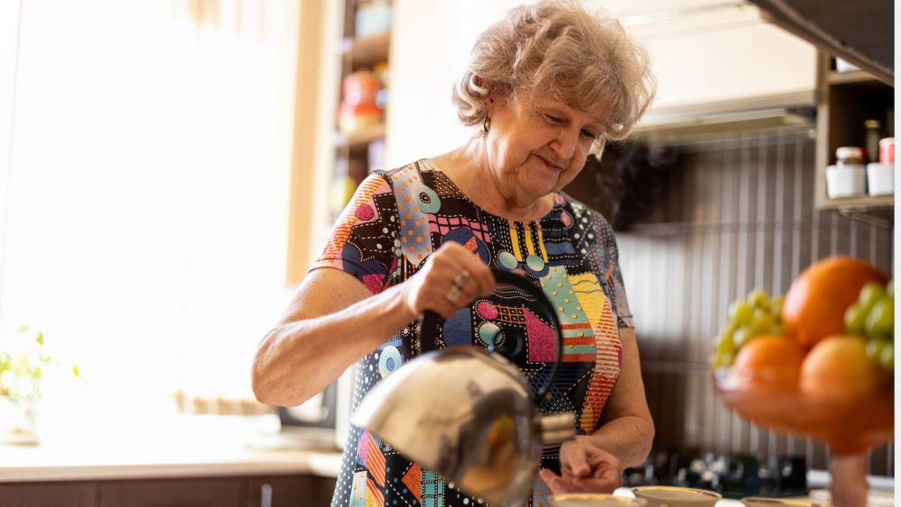 Image of lady with grey hair and multi-coloured top making a cup of tea in her kitchen.