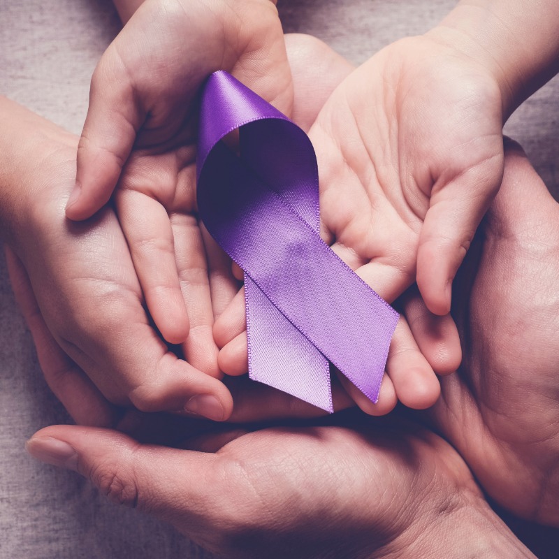 Image of three sets of hands on top of each other holding a purple ribbon which represents domestic abuse awareness.
